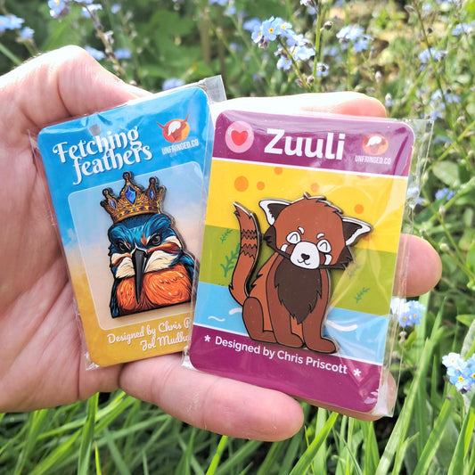 Two pin badges featuring 'Fetching Feathers' and 'Zuuli' against a natural background with flowers and grass.